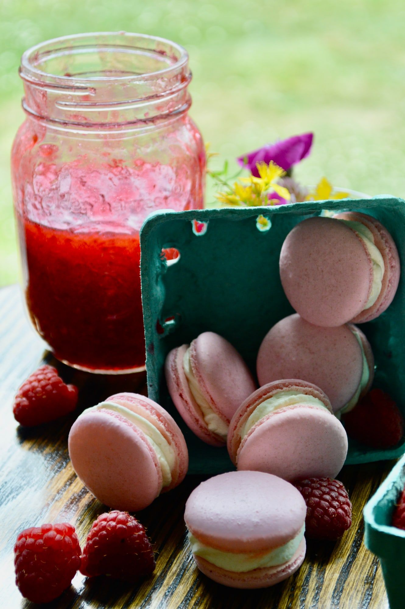 Pink macarons in a berry carton beside a jar of raspberry jam and fresh berries