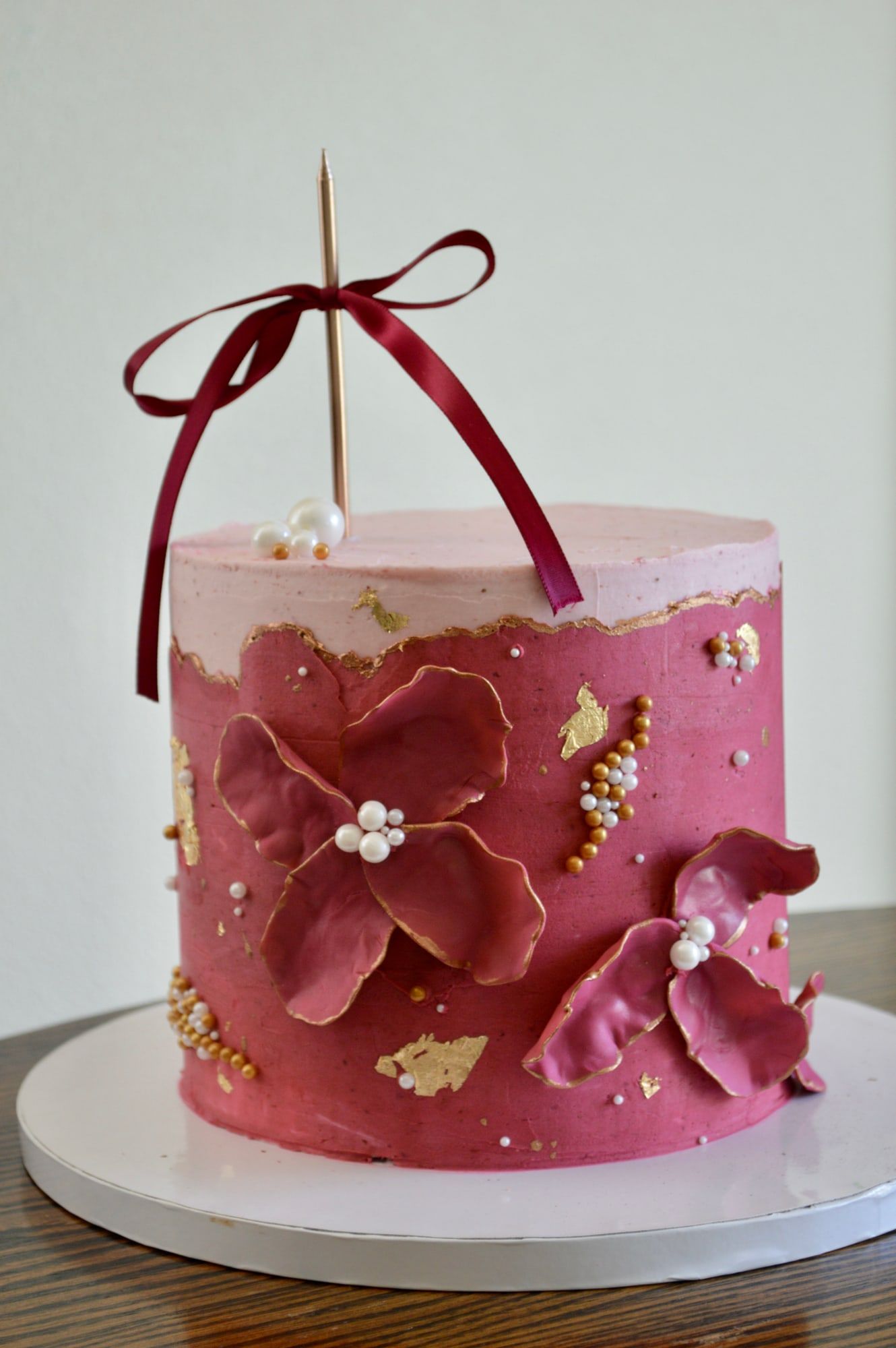 Pink cake with burgundy sugar flowers, gold leaf and a ribbon topper