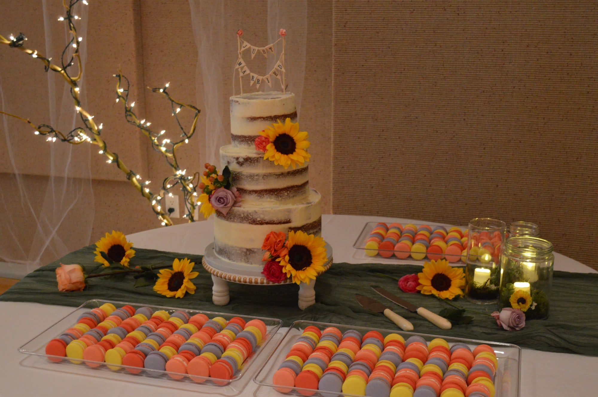 Semi-naked sunflower wedding cake on a lit dessert table beside trays of macarons